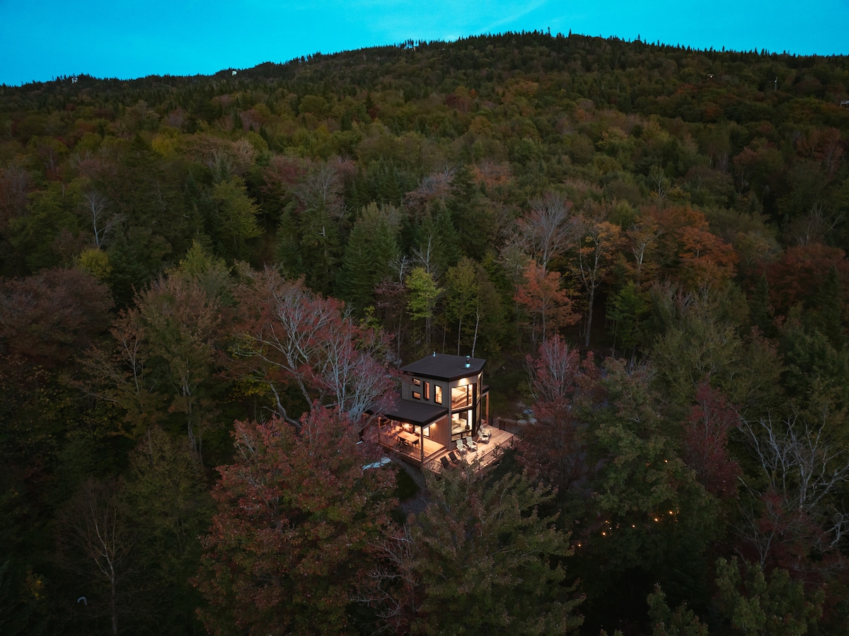 The micro-chalet is nestled among vibrant autumn trees, surrounded by a natural landscape. The structure features illuminated outdoor areas, enhancing visibility at dusk. The backdrop of the rolling hills offers a serene and tranquil setting, highlighting the chalet's connection to nature.
