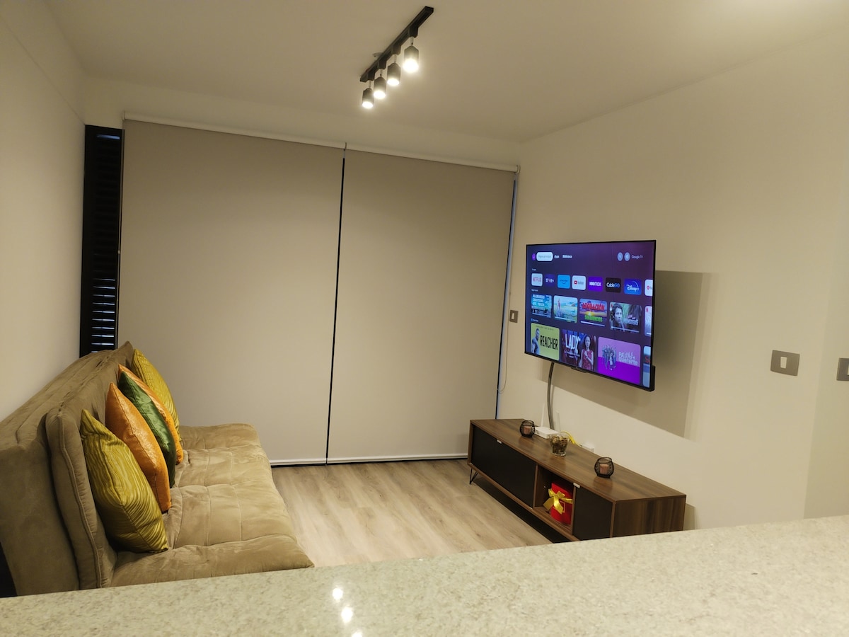 A living area features a beige sofa adorned with colorful cushions and a sleek media console. A wall-mounted television displays streaming options, and natural light is filtered through roller shades, complemented by wooden flooring that adds warmth to the space.