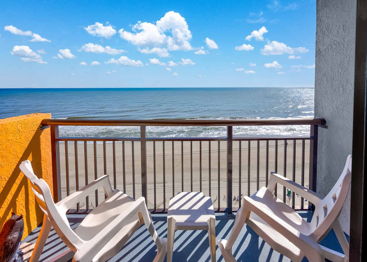 A private balcony features two white lounge chairs and a small round table, positioned to overlook the sandy beach and ocean. The vibrant blue sky is dotted with fluffy white clouds, providing a serene backdrop to the sparkling water and shoreline.