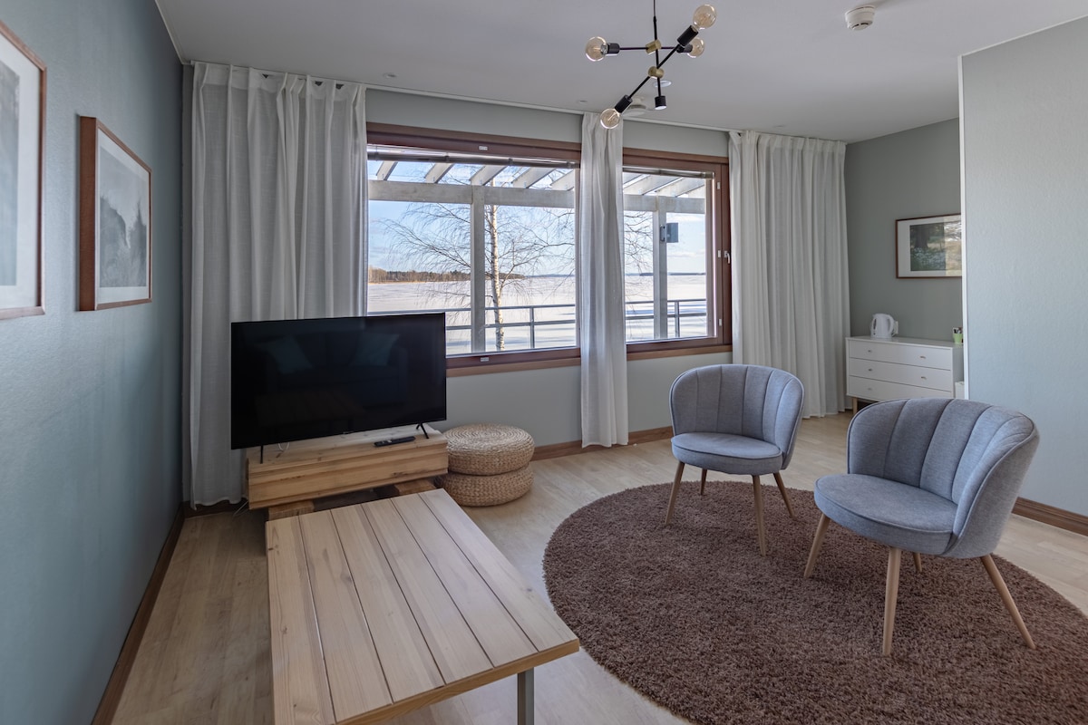 A light-filled living area features a large window with sheer curtains and a balcony view. Two light blue armchairs are positioned nearby a wooden table, resting on a circular rug. A wall-mounted television and a sideboard add to the minimalistic decor.