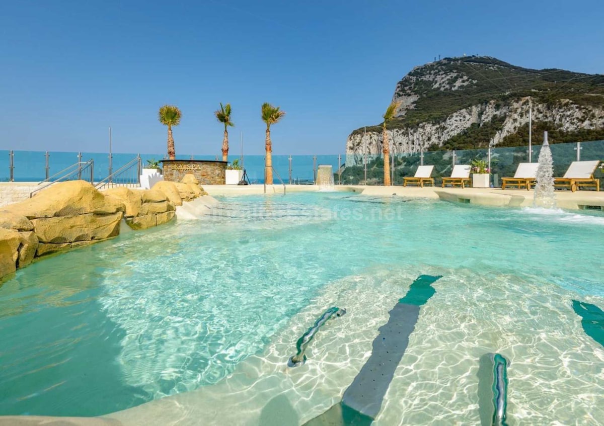 A serene pool area offers inviting water features, surrounded by comfortable sun loungers and palm trees. Clear blue skies provide the backdrop for the picturesque scenery, and a rocky hillside is visible in the distance, enhancing the tranquil atmosphere.