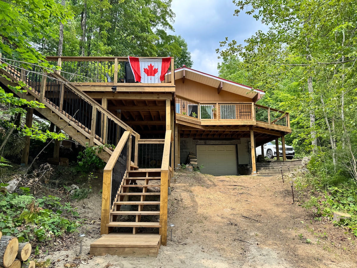The exterior of the cottage showcases a wood deck with a Canadian flag displayed prominently. A set of wooden steps leads to the entrance, surrounded by greenery. The structure is elevated with ample space underneath, complemented by a dirt area and a gravel parking space nearby.