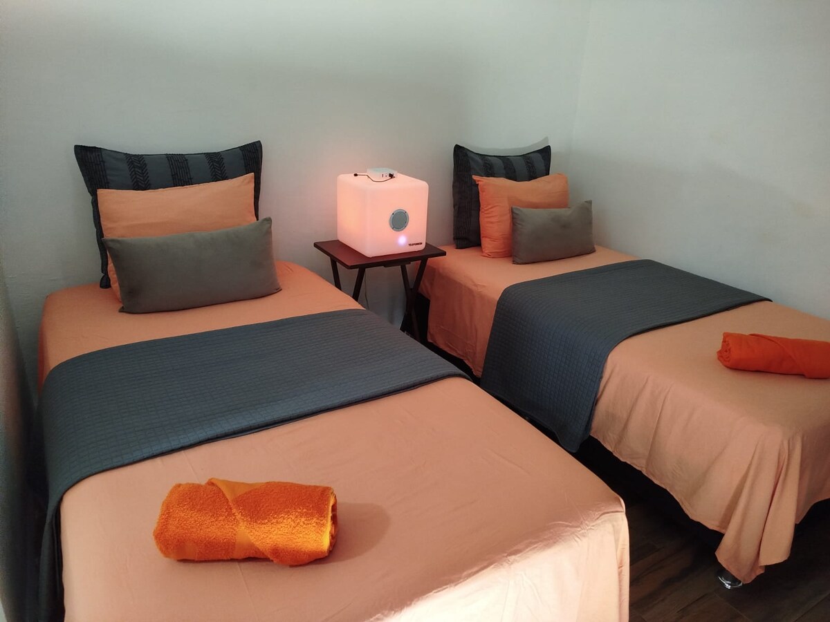 The image depicts a serene sleeping area featuring two individual beds dressed in soft peach linens. Each bed is separated by a small table with a light source. Accent pillows in varying shades of gray and orange are arranged atop the beds.