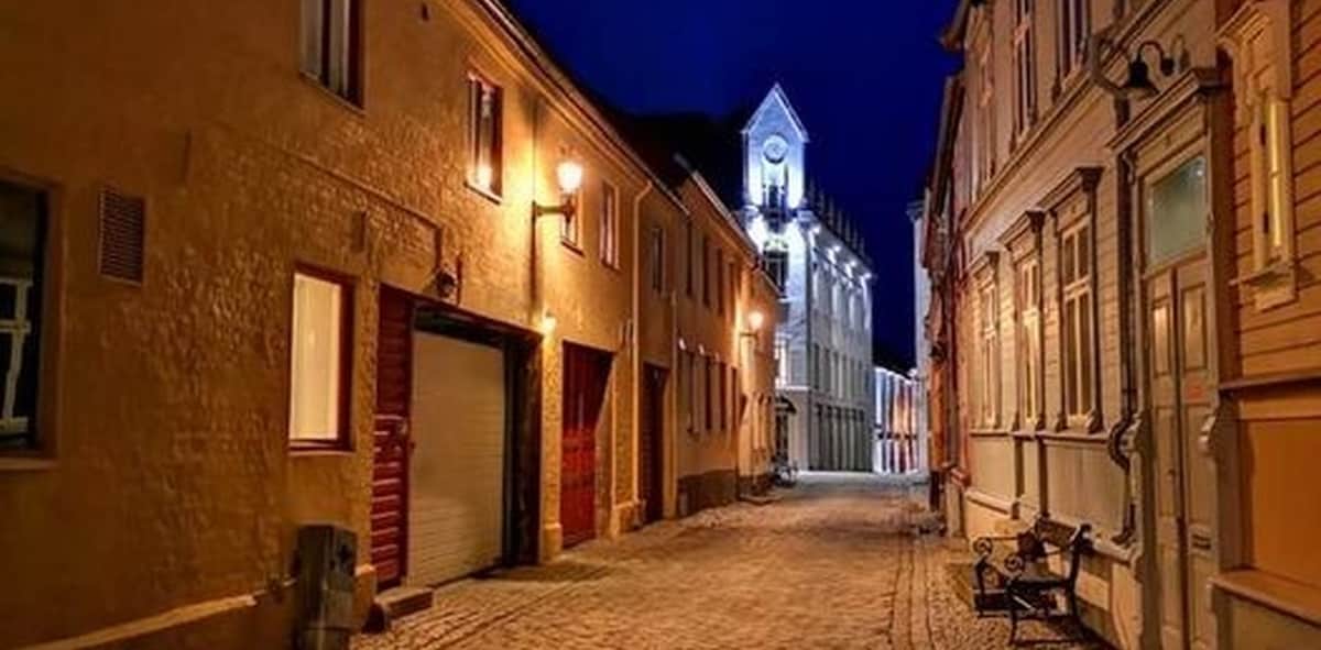 A quiet cobblestone street is illuminated by warm street lamps at night. The charming architecture features historic buildings with varying window styles and façades, leading toward a well-lit structure at the end of the street. Benches are visible along the walkway, inviting relaxation.