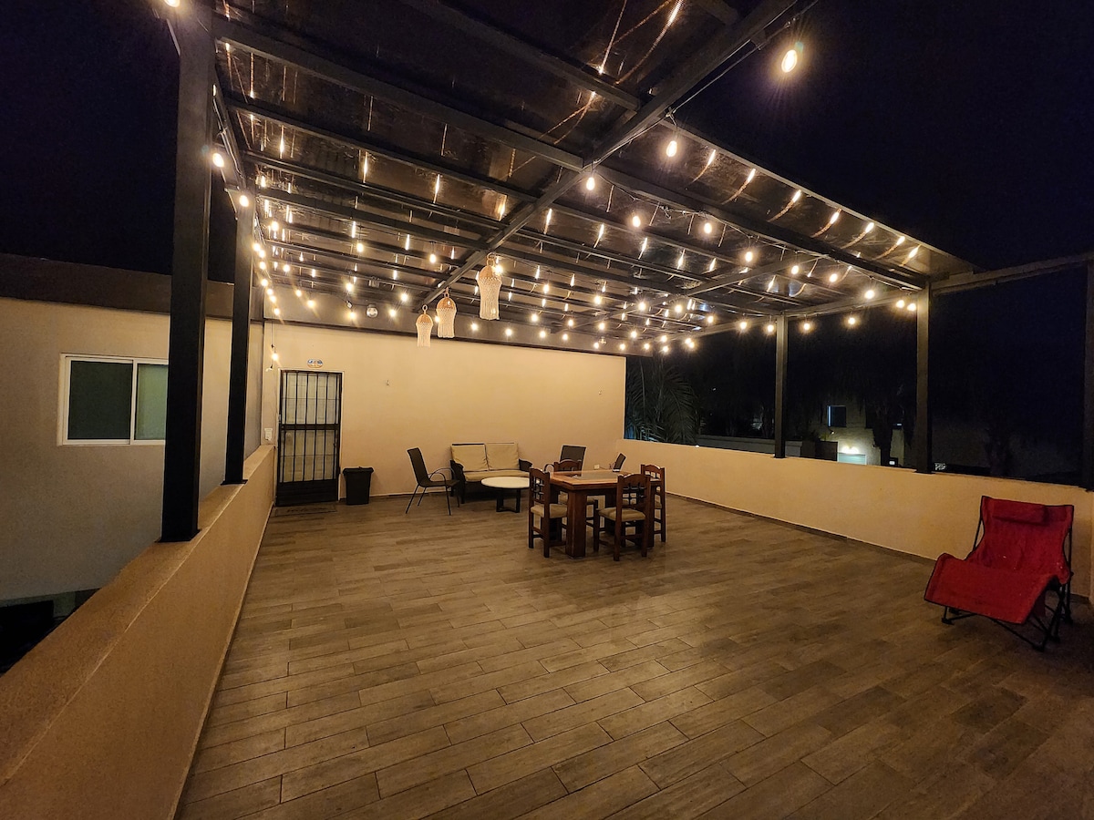 An expansive terrace is illuminated by string lights creating a serene ambiance at night. Multiple seating arrangements are present, including a large wooden table surrounded by chairs. A pair of lounge chairs is positioned for relaxation. The space is covered, offering protection from the elements.
