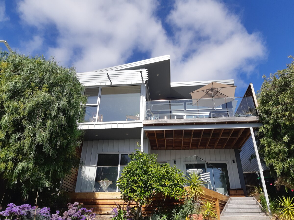 A contemporary home features large glass windows and a spacious deck. An umbrella provides shade over the outdoor seating area, while lush greenery and vibrant flowers surround the entrance, enhancing the inviting appearance of the property.