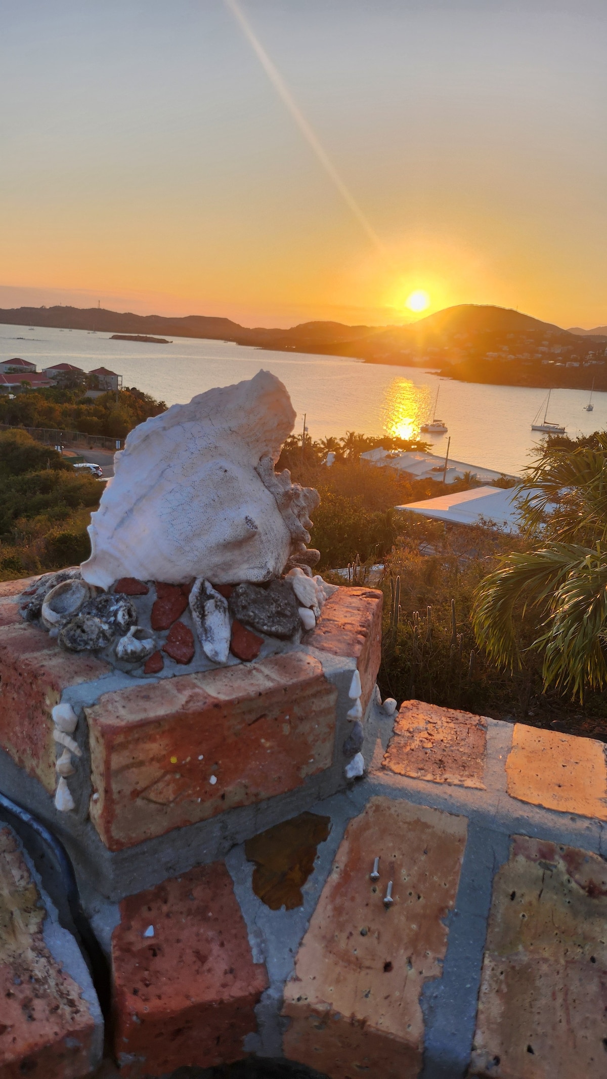 A decorative arrangement of seashells rests atop a brick structure, with the sun setting over the tranquil water in the background. The horizon blends warm hues of orange and yellow, illuminating the landscape and creating a peaceful ambiance.