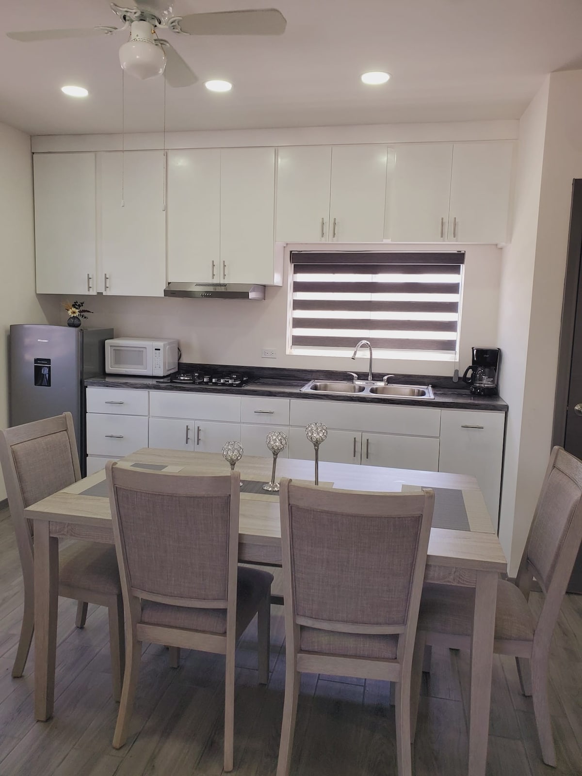 A modern kitchen equipped with essential appliances is presented. The space includes a stainless steel refrigerator, a microwave, and a stove. A dining table with four chairs is arranged nearby, complemented by a decorative centerpiece. Natural light is filtered through dual-tone window shades.