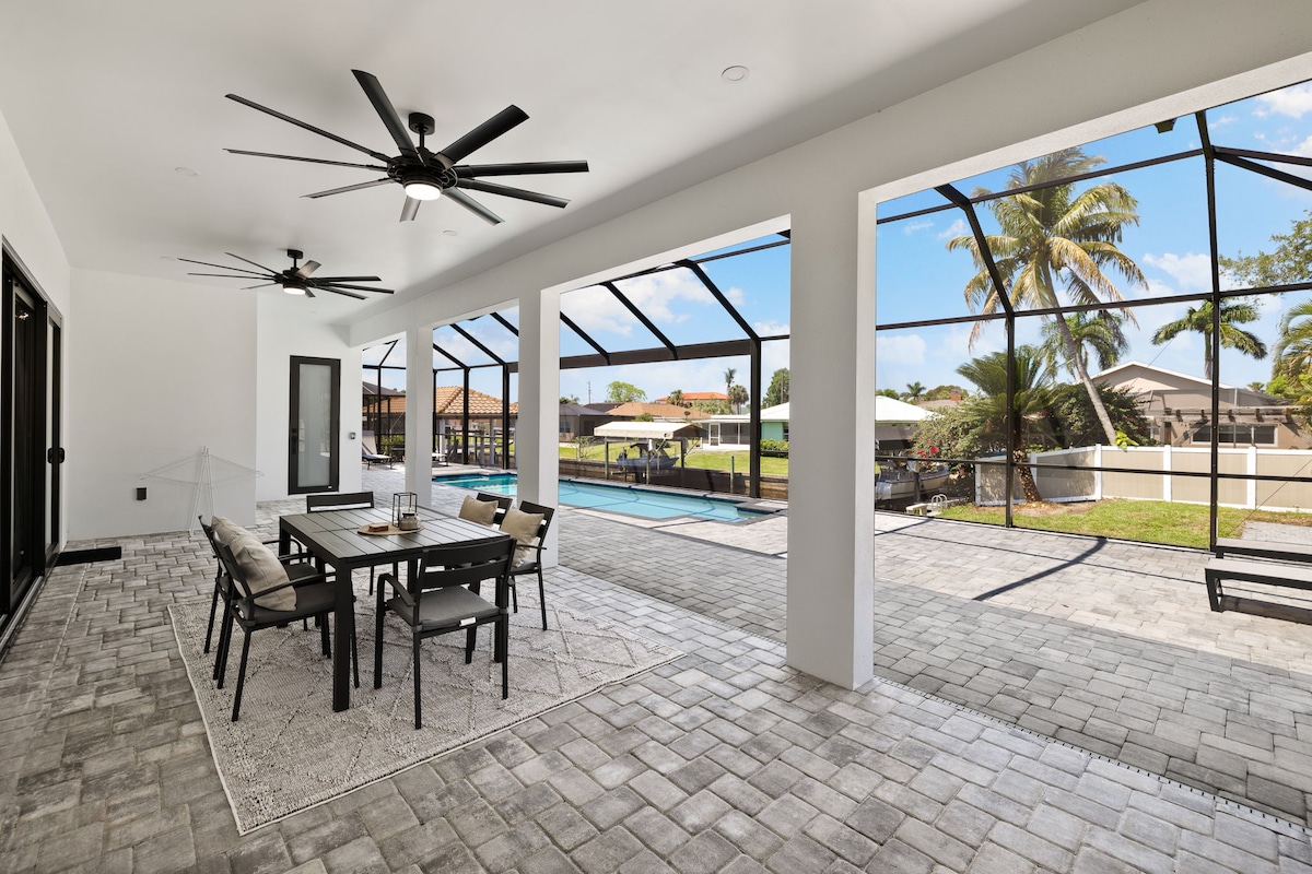 An open-air patio features a dining table surrounded by chairs, offering a serene space. Two ceiling fans are visible above. Large screens line the walls, providing views of the outdoor pool and landscaping, while ample light filters through the clear openings.