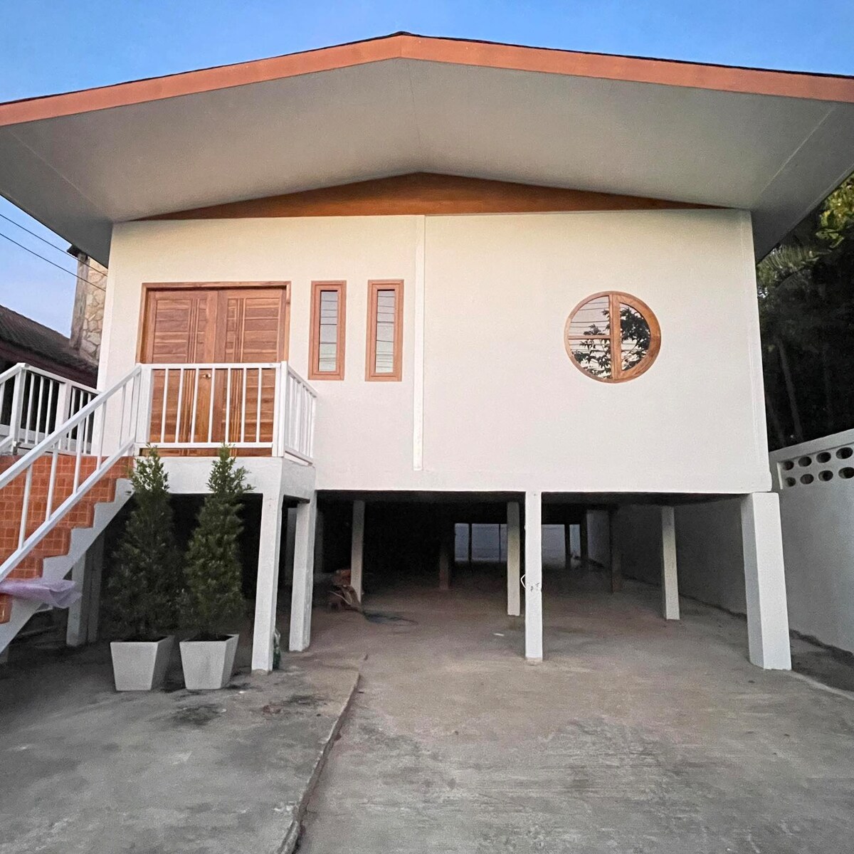The exterior of the house displays a raised structure with a flat roof. Two wooden doors are visible, accompanied by a large circular window. A driveway leads to an open space beneath the house, with two potted plants on either side of the entrance.