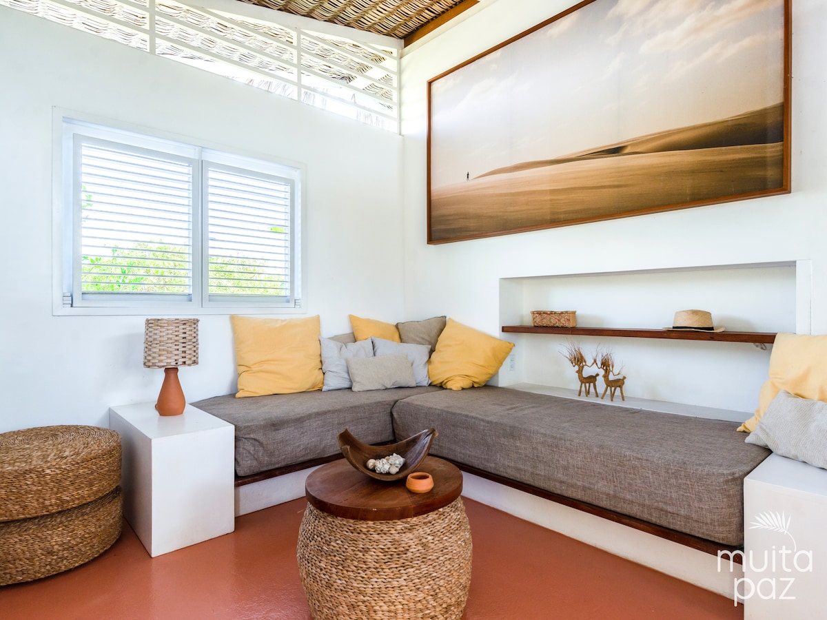 A cozy seating area features a sectional sofa adorned with soft yellow and neutral cushions. A rustic wooden table sits in the center, complemented by woven accents. Light pours in through a window with horizontal blinds, enhancing the serene ambiance.