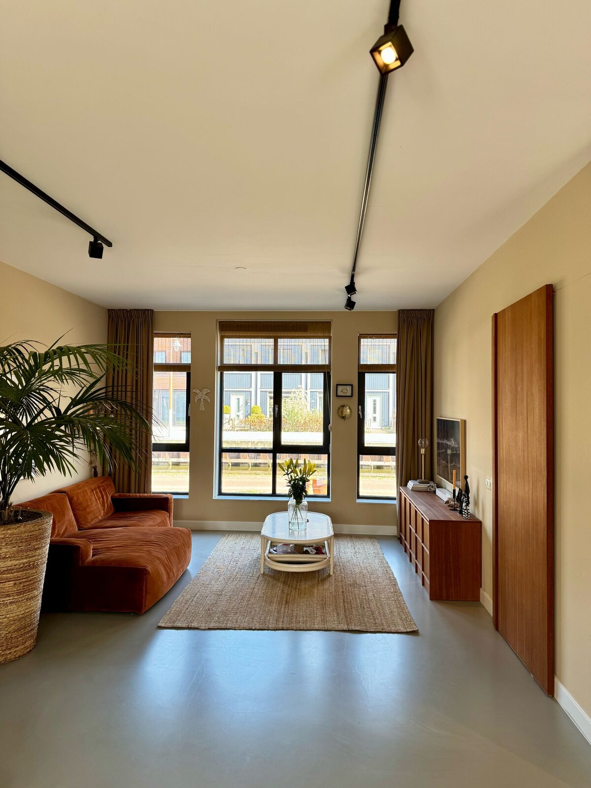 A spacious living area is presented featuring large windows that allow natural light to fill the room. A comfortable sofa and minimalist coffee table provide seating and a central gathering space. Decorative plants add a touch of greenery, enhancing the inviting atmosphere.