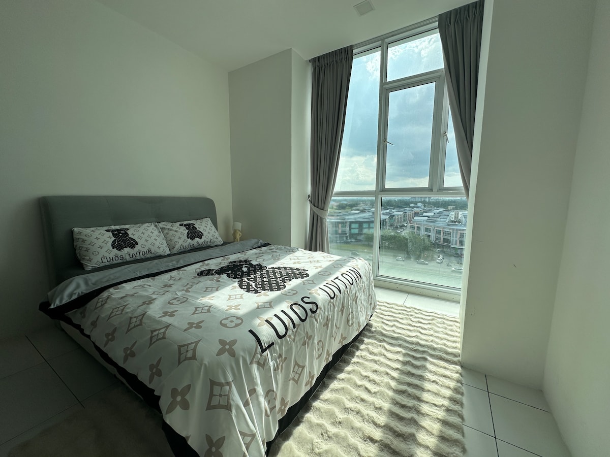 A bright bedroom features a spacious bed covered with a patterned duvet and matching pillows. Natural light pours in through a large window, creating a warm ambiance. Light, neutral tones are complemented by soft, textured rugs on the floor.