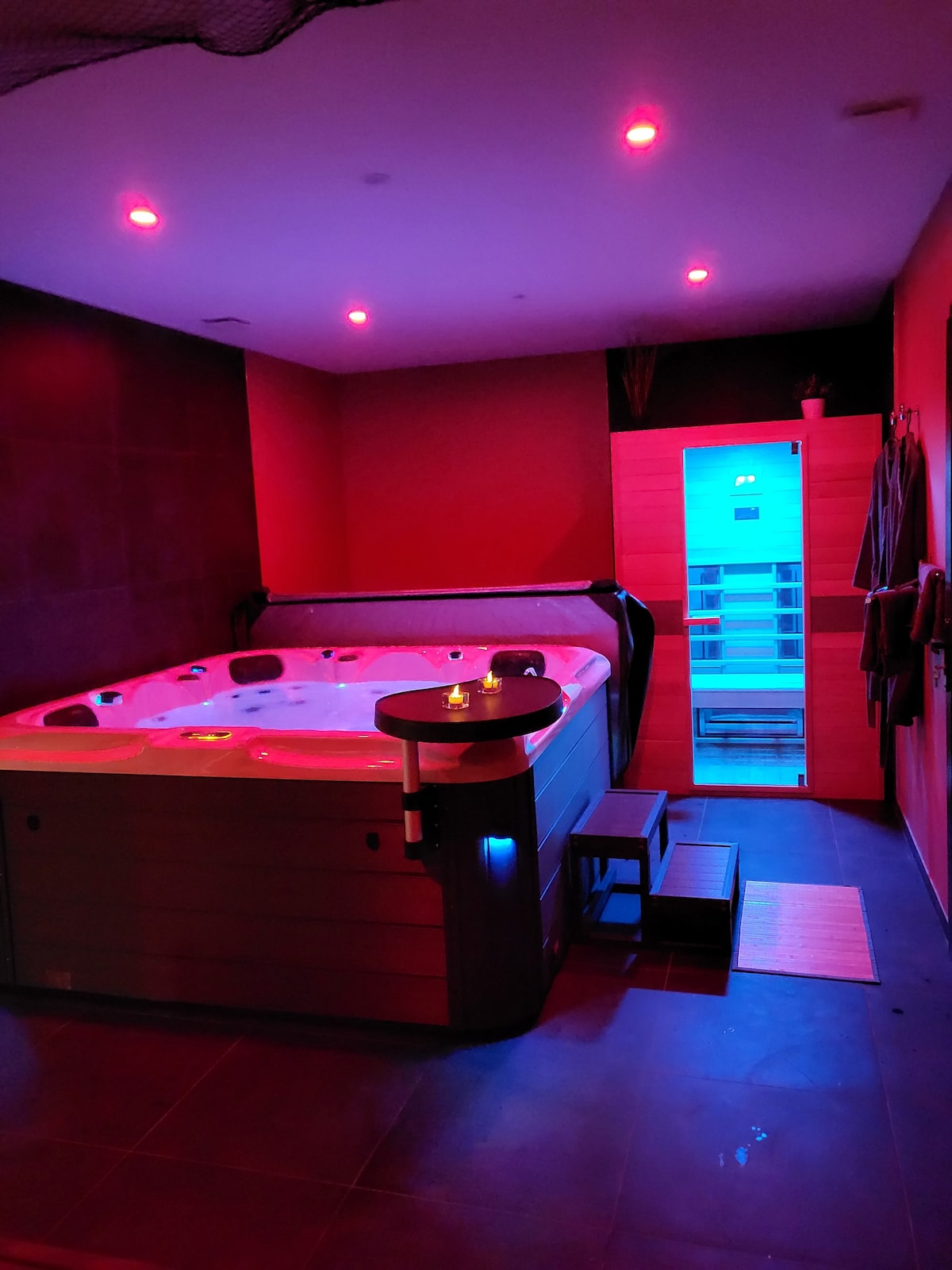 An inviting relaxation area features a spacious jacuzzi, illuminated by soft red lighting. A sauna is visible in the background, emitting a calming blue hue. Subtle decor elements enhance the tranquil atmosphere, while a few steps lead up to the jacuzzi for easy access.