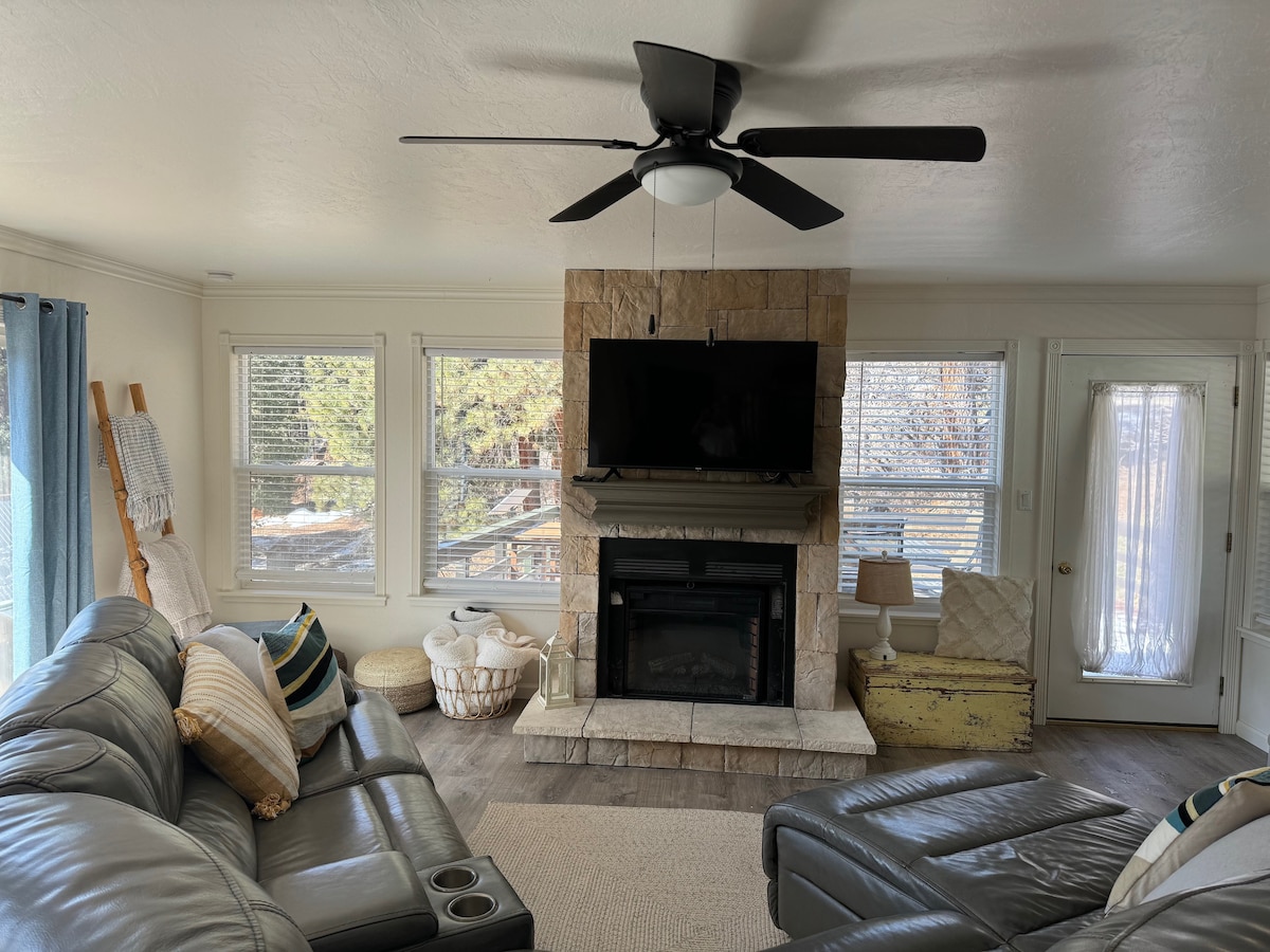 A spacious living room features a stone fireplace as the focal point, with a large flat-screen television mounted above. Two leather sofas, accented by colorful throw pillows, are arranged to encourage relaxation. Natural light filters through several windows, illuminating the cozy interior.