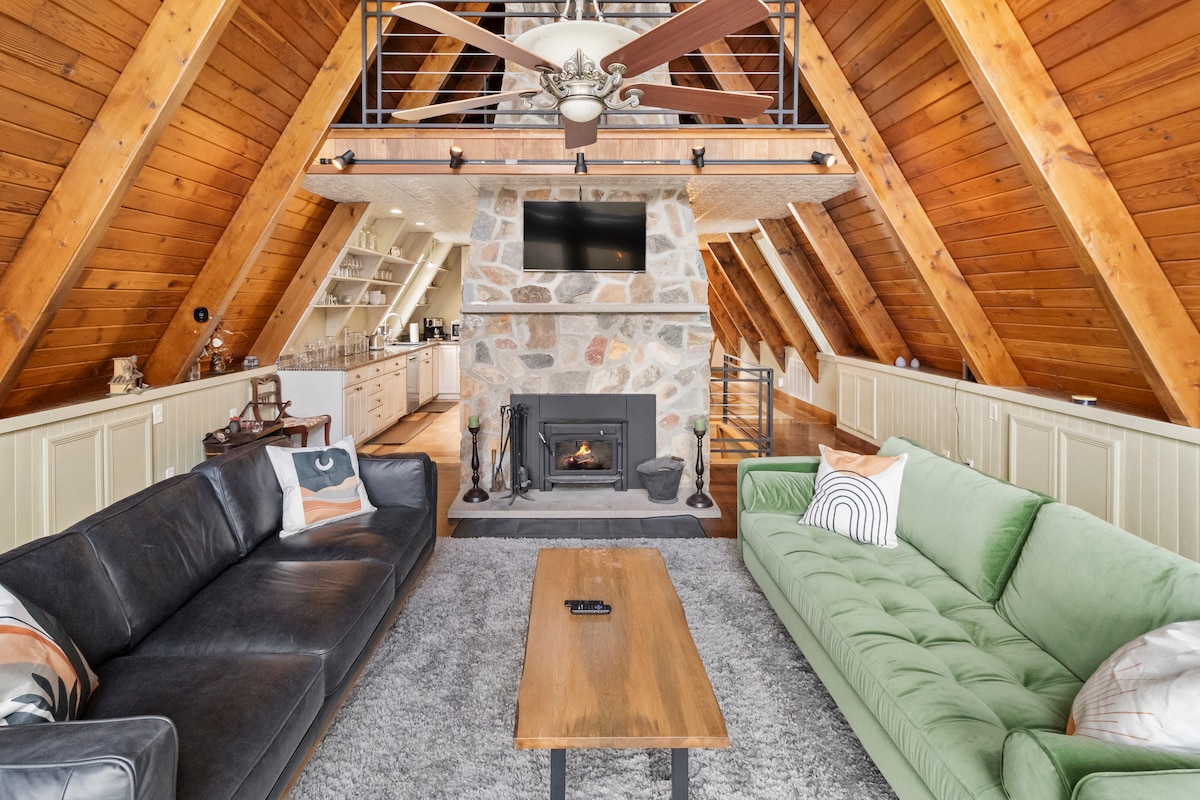 The open living area features high, wooden A-frame ceilings with exposed beams. A large stone fireplace is centered between two inviting sofas—one dark and the other green. A wooden coffee table sits in front of the sofas, with a wall-mounted television above the fireplace.