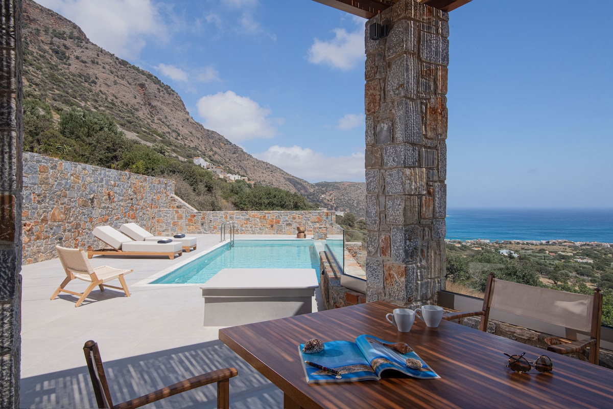 An outdoor dining area features a wooden table set with cups and a magazine. Loungers are positioned beside a swimming pool, with views extending towards the mountains and the sea. Bright blue skies complement the serene landscape.