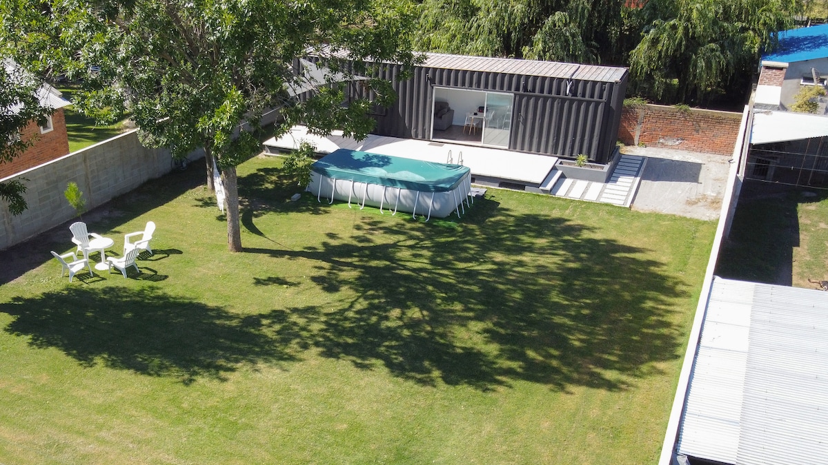 A spacious outdoor area is surrounded by greenery, featuring a large lawn with a tree providing shade. A swimming pool is visible alongside a modern structure with large windows. Two chairs are arranged nearby, inviting relaxation in the open air.