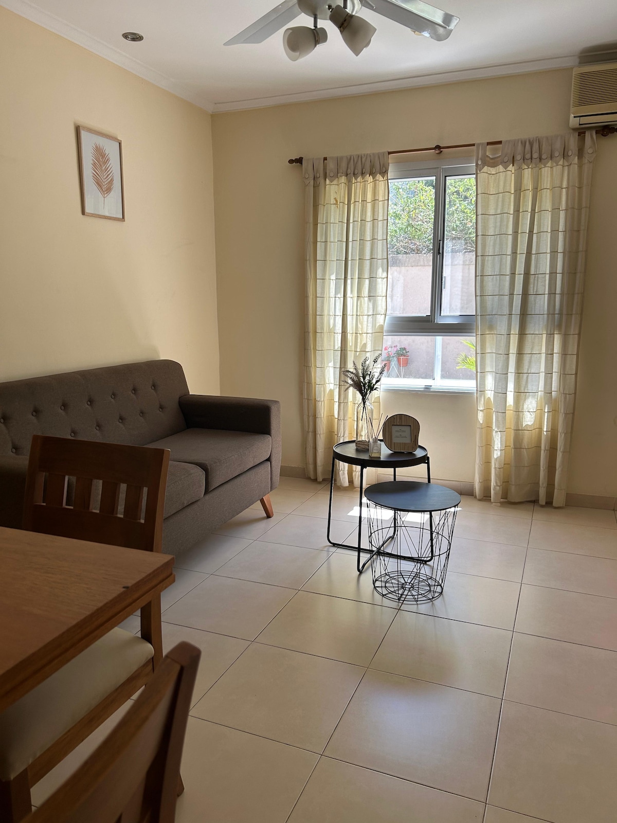 A well-lit living area is featured, showing a gray sofa next to a small round table. A window, partially covered by light curtains, allows natural light to fill the room. A dining table and chairs are present, enhancing the space's functionality.