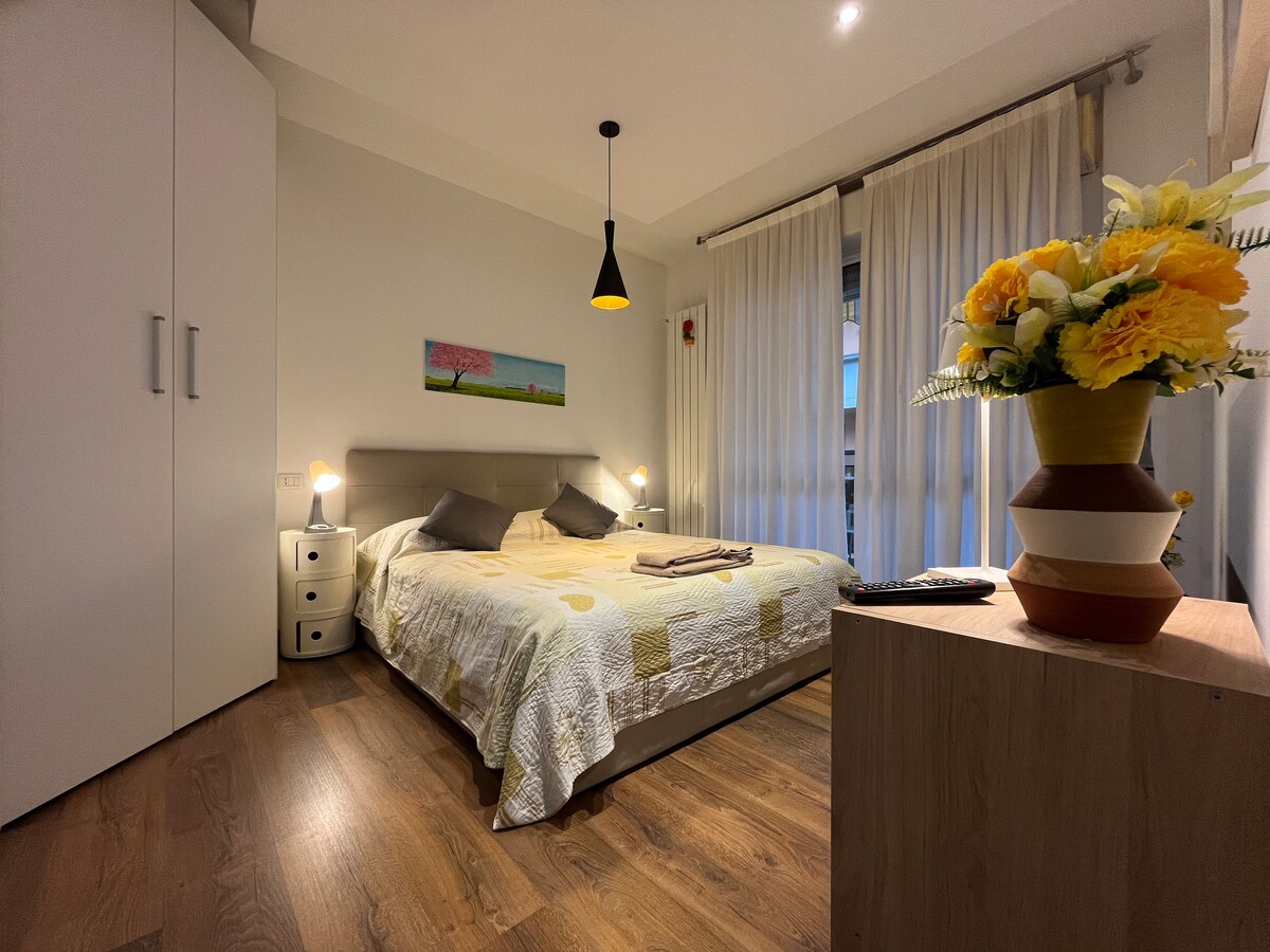 A well-lit bedroom features a spacious bed with light-colored bedding. Soft gray pillows are neatly arranged, and bedside tables with lamps are placed on either side. A vibrant painting is displayed above the bed, and sheer curtains allow natural light to fill the room.