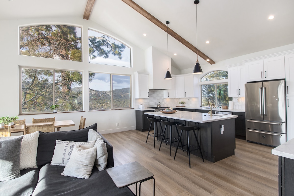 A modern kitchen is illuminated by natural light from large windows, offering mountain views. White cabinetry contrasts with dark countertops and stainless steel appliances. A spacious dining area features a table and chairs, while a comfortable sofa is positioned in the open living space.