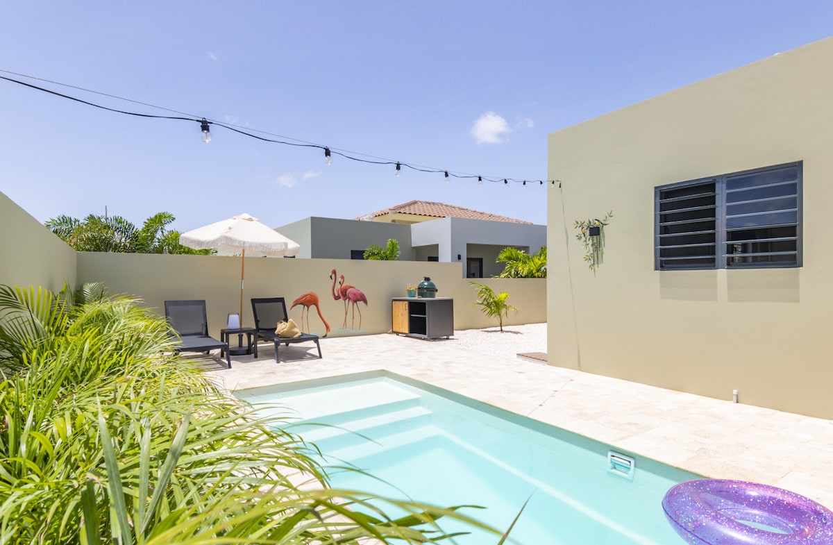 A private outdoor space features a turquoise pool surrounded by beige stone tiles. Two lounge chairs and a small table are positioned under an umbrella. Colorful flamingo murals decorate the walls, complemented by lush greenery.