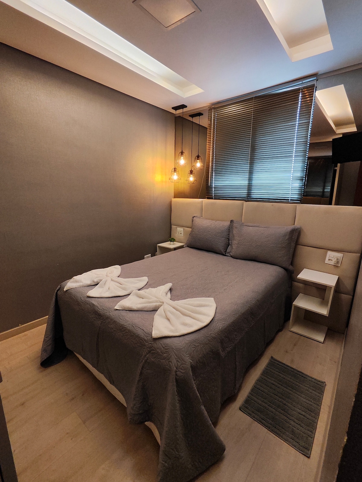 A sleeping area features a spacious bed adorned with neatly arranged towels. Soft lighting is provided by pendant bulbs above the bed. Window blinds allow for light control, while a small nightstand stands beside the bed, against a muted wall.