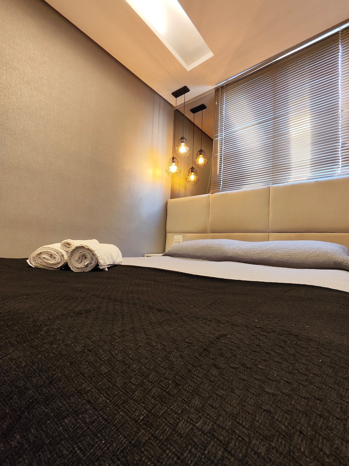 A cozy bedroom features a spacious bed with dark bedding. Neatly rolled towels are placed at the foot of the bed. Soft lighting is provided by pendant bulbs hanging above, while natural light filters through the window with horizontal blinds.