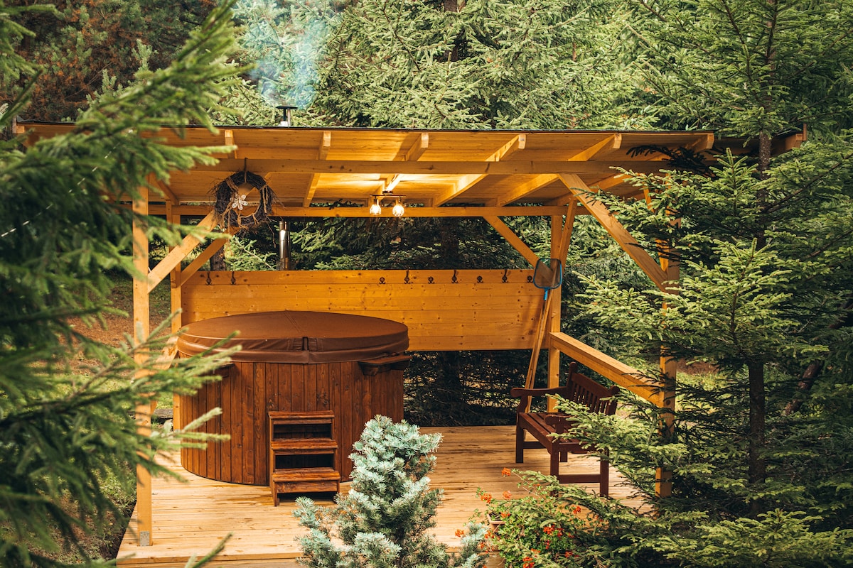 A wooden hot tub is situated under a canopy structure, surrounded by tall trees and greenery. A small wooden bench is positioned nearby, while soft lighting highlights the warm wood tones of the shelter. Natural elements create a serene, private setting.