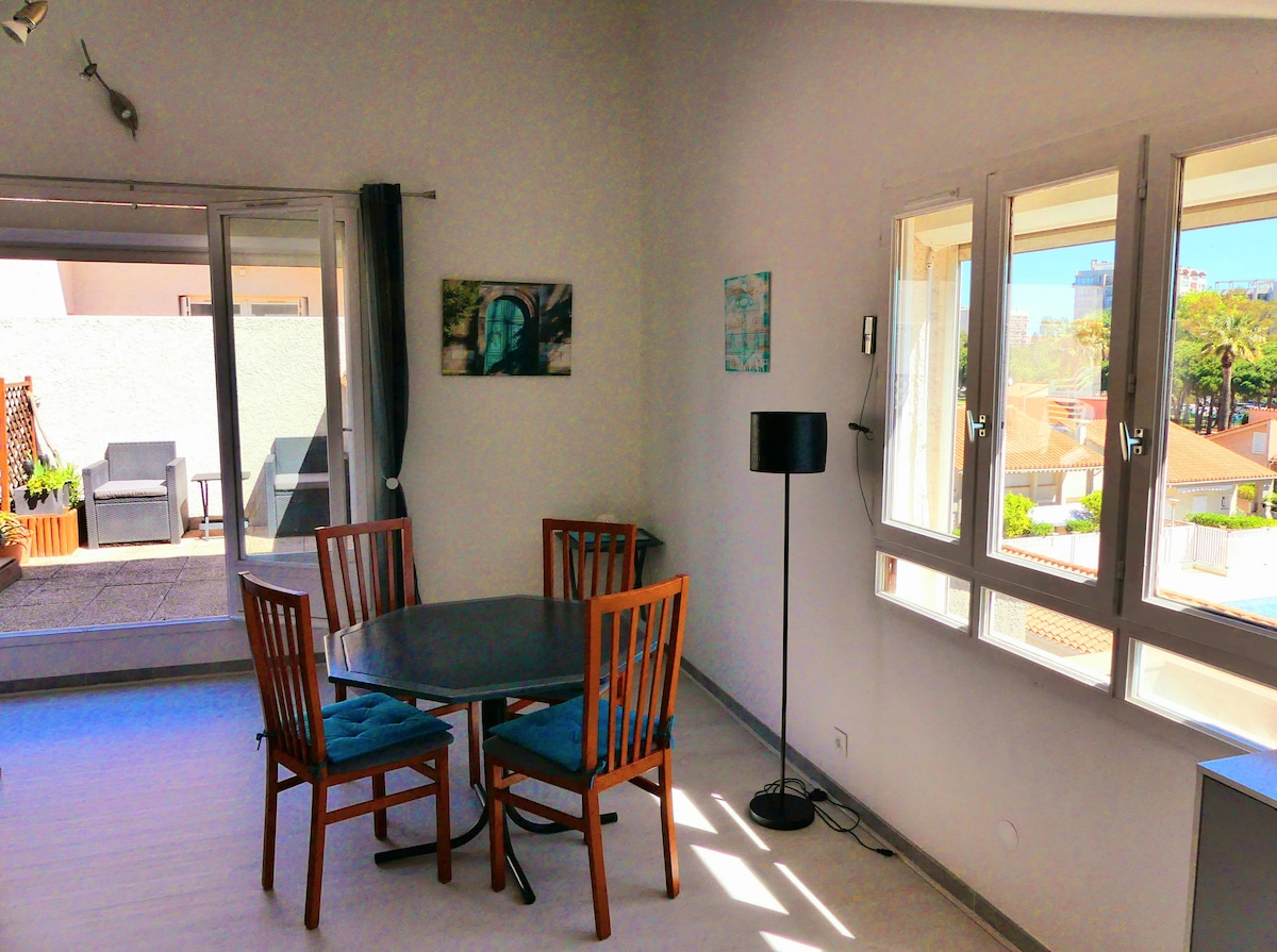A spacious dining area features a round table with four wooden chairs and a floor lamp. Natural light enters through large windows, providing a view of the terrace and surrounding greenery. A piece of wall art complements the bright, neutral walls.