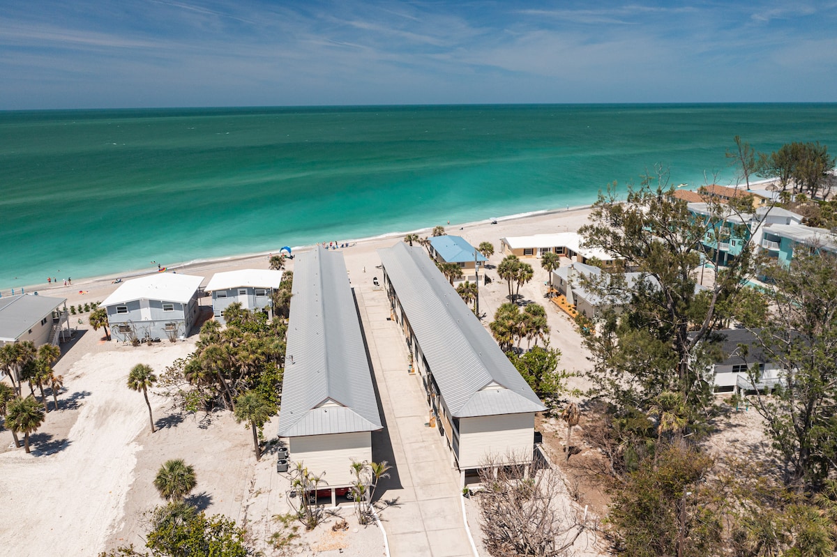 An aerial view captures the layout of the condo complex bordered by lush palm trees and sandy paths. The turquoise waters of the beach stretch beyond, with a clear sky overhead. The proximity to the shoreline invites outdoor enjoyment and leisure activities.