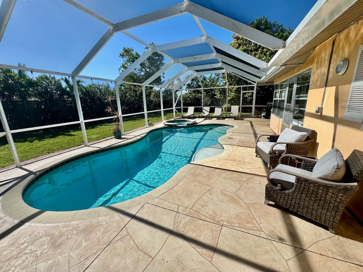 A heated pool is surrounded by a screened lanai, providing a bug-free zone. Outdoor loungers are positioned nearby, and a cozy seating area offers plush chairs. Lush greenery is visible in the background, ensuring privacy and a tropical feel.