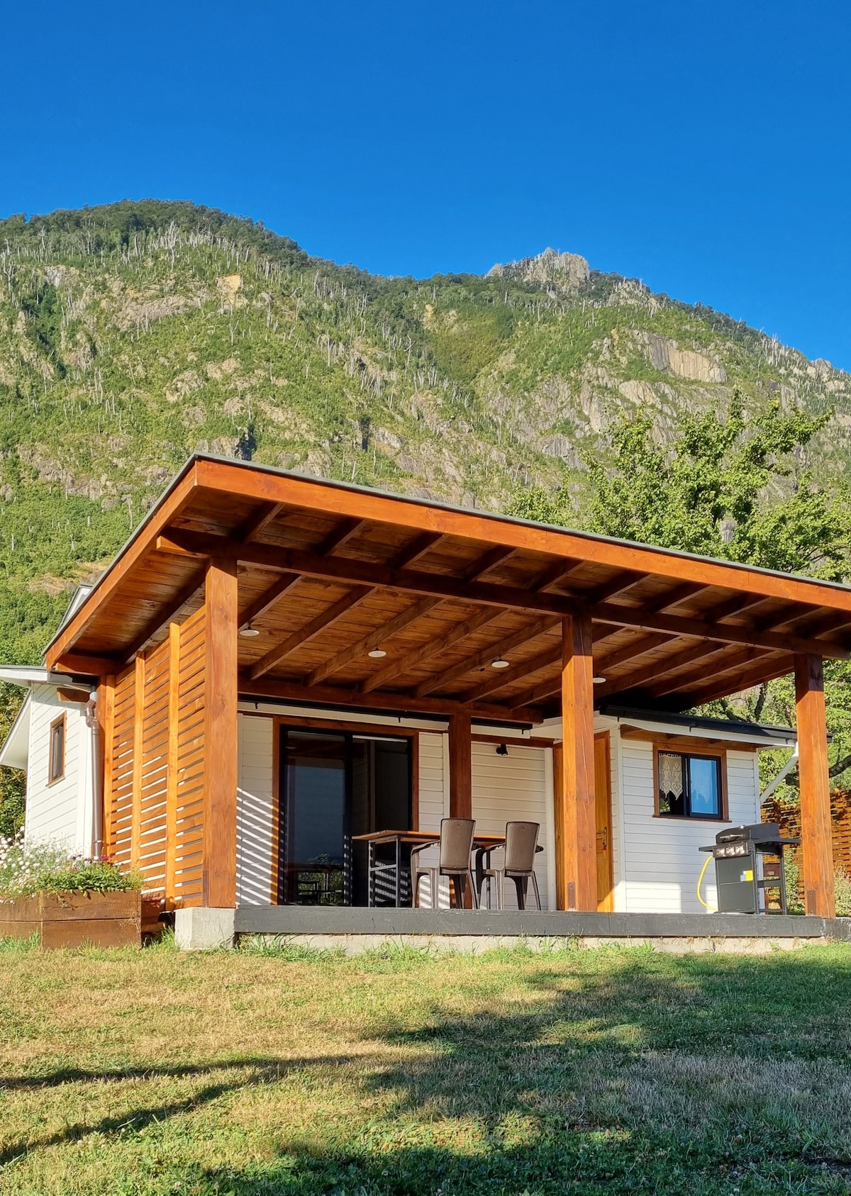 The cabin exterior is framed by lush greenery and towering mountains in the background. A covered porch features a small dining table with four chairs and a gas grill, providing a welcoming space for outdoor relaxation.