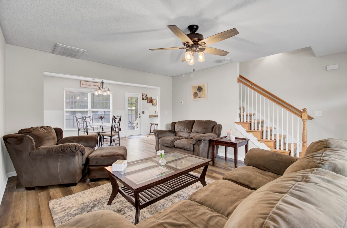 A spacious living area features comfortable seating arranged around a coffee table. A ceiling fan provides airflow, while light streams in through multiple windows. In the background, a dining area can be seen, with a staircase leading to the upper level.