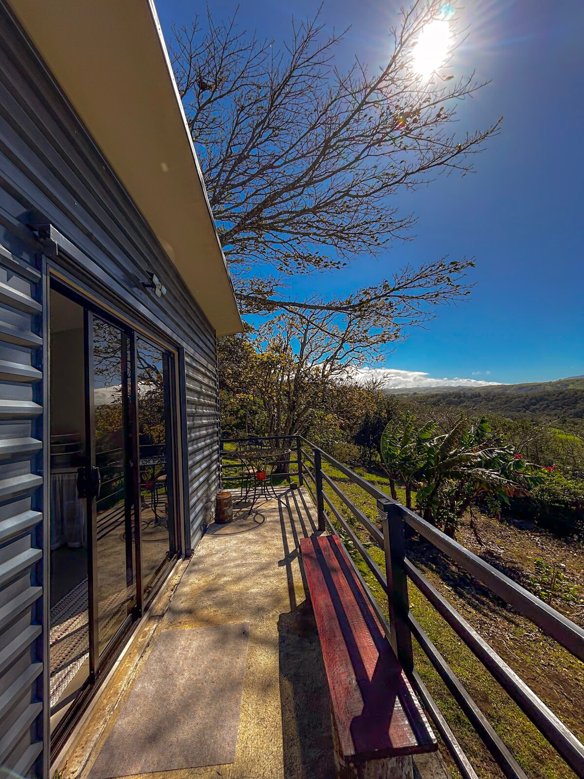 A spacious deck extends from the cabin, offering views of lush greenery and distant hills. Large sliding glass doors provide access to the interior, while a red bench sits along the railing. The bright sun illuminates the area, enhancing the outdoor experience.