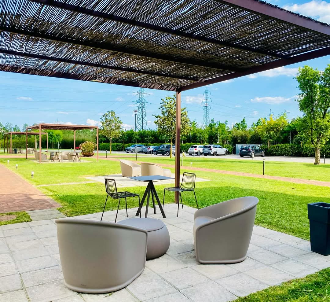 An outdoor seating area is shaded by a pergola, featuring modern chairs and a round table atop a stone patio. Lush grass surrounds the space, with trees and parked cars visible in the distance under a clear sky.