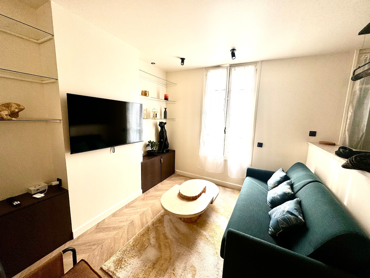 A contemporary living area features a green sofa and a stylish low coffee table. A large TV is mounted on the wall, accompanied by shelves displaying decorative items. Natural light enters through two windows dressed in sheer curtains, enhancing the overall ambiance.