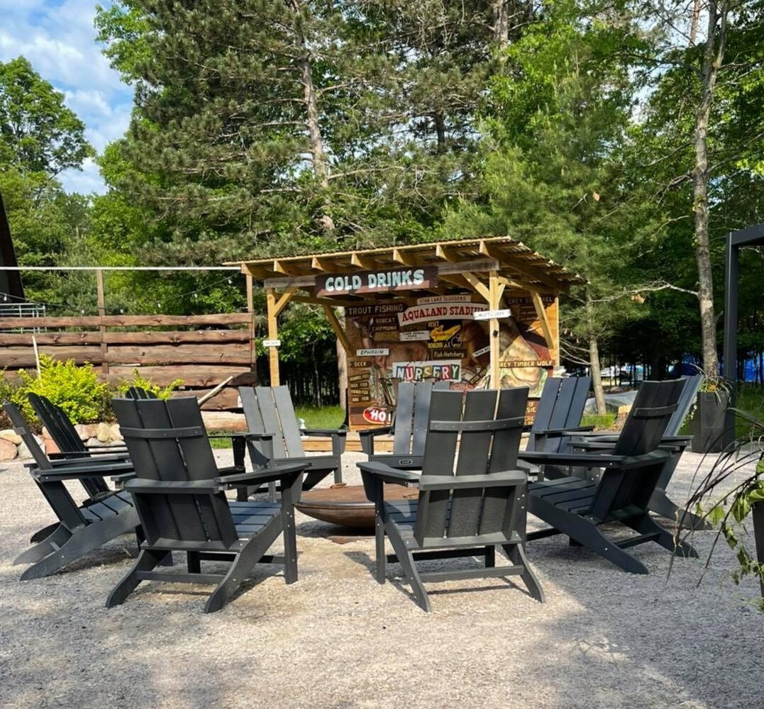 A gathering area is surrounded by several dark wooden Adirondack chairs arranged in a circle around a fire pit. A shaded structure displays the words 'COLD DRINKS' and offers a scenic backdrop of trees, inviting outdoor relaxation.