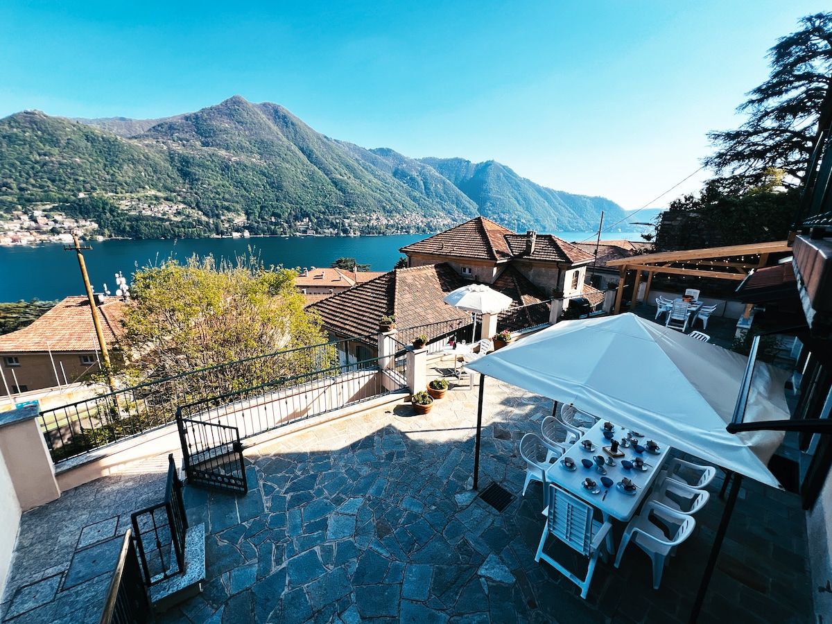 A spacious outdoor terrace is presented, featuring a dining table set for meals and a large umbrella for shade. Scenic views of Lake Como and surrounding mountains are visible, while terracotta roofs of nearby buildings are arranged in the background against a clear blue sky.