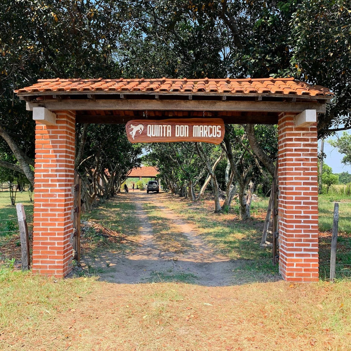 A brick entrance with a clay tile roof welcomes guests to Quinta Don Marcos. The pathway is lined with trees on both sides, leading to an open area beyond. A parked vehicle is visible in the distance, indicating accessibility and ease of arrival.