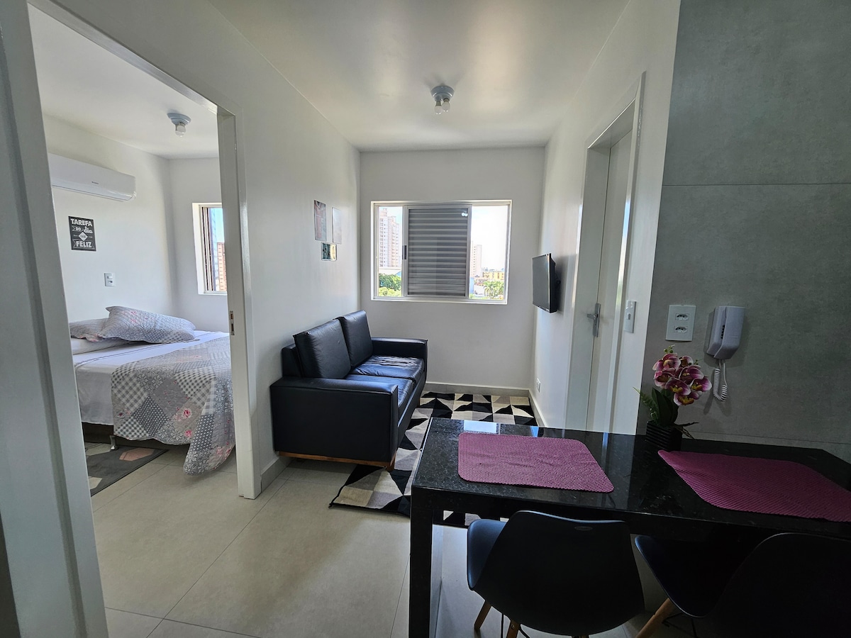 A bright and welcoming living area features a black sofa paired with a modern coffee table. A compact dining space is equipped with chairs and a table setting. The separated sleeping area includes a cozy bed, and daylight filters through the window enhancing the open layout.
