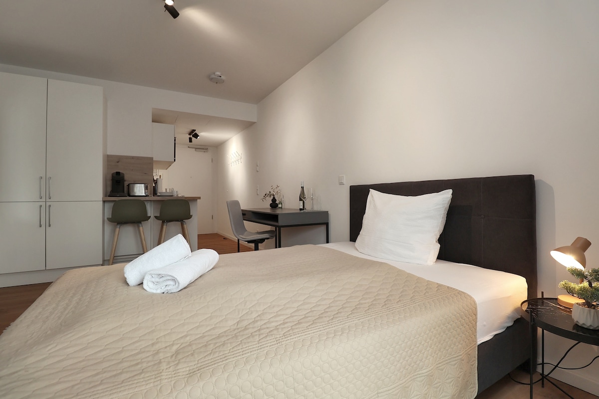 A well-appointed studio space features a comfortable bed dressed in neutral tones. The kitchen area is visible in the background, equipped with modern cabinetry and appliances. A dining table and chair are present, providing a place for meals or work, while natural light softly fills the room.