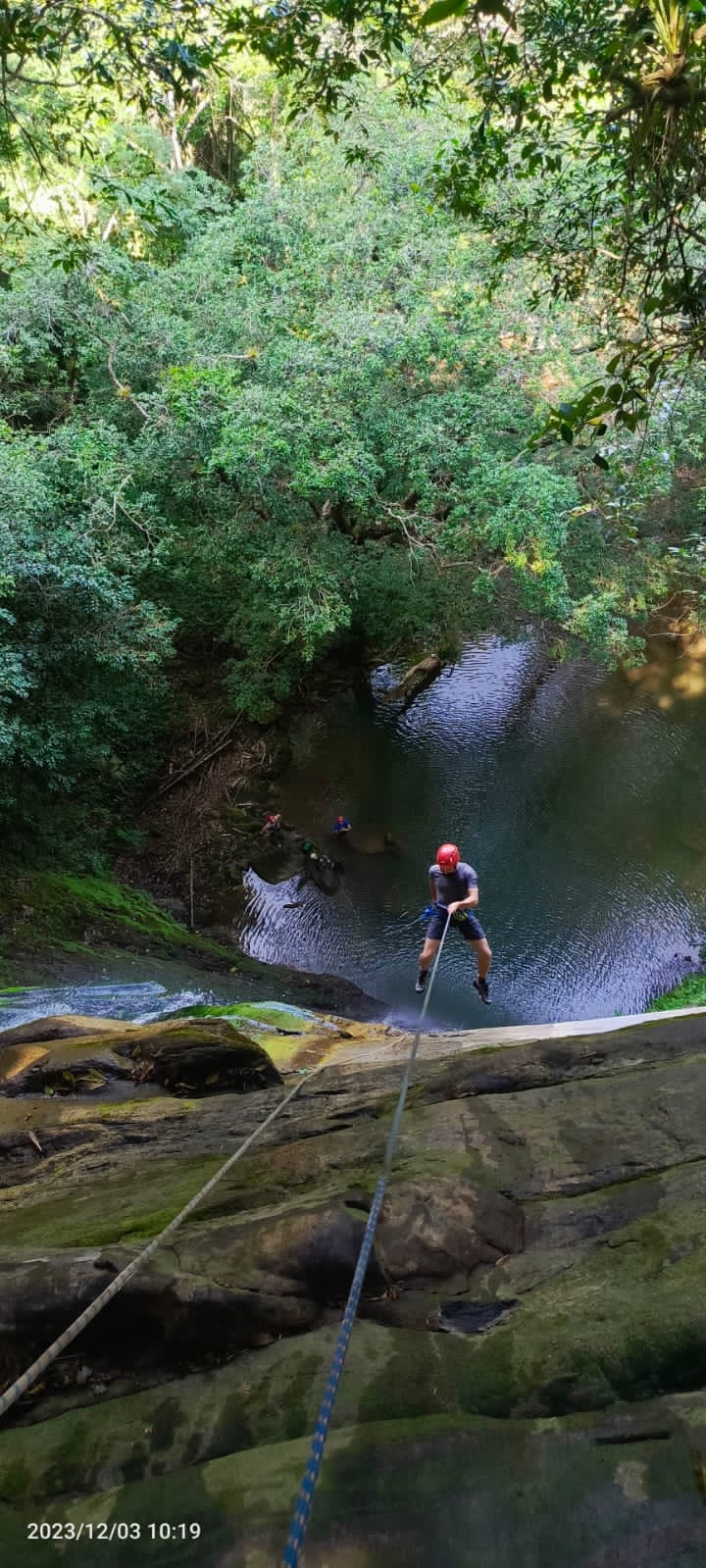 A person is seen descending a rocky cliff with a harness and rope. Below, a serene waterbody is surrounded by lush greenery, with two individuals partially visible in the water. The scene captures a blend of adventure and natural beauty.