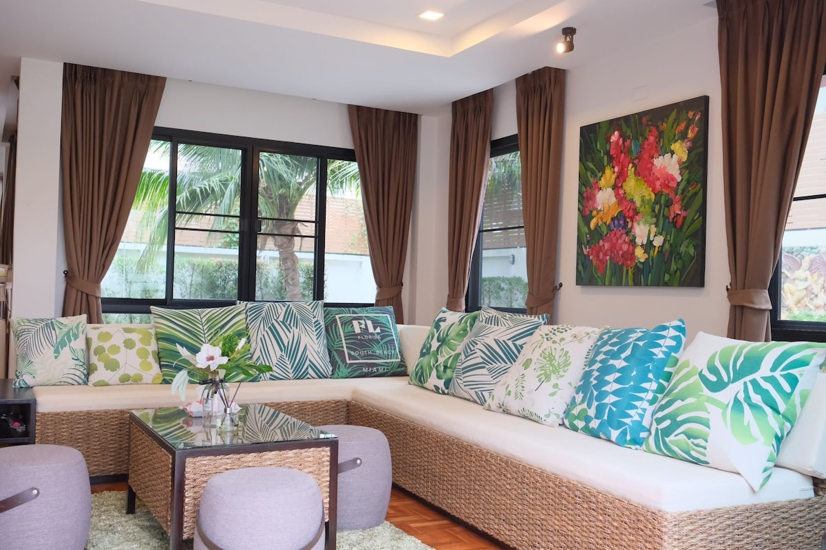A spacious living area features large windows framed by soft brown curtains, allowing natural light to illuminate the room. A sectional sofa adorned with colorful tropical-patterned cushions surrounds a glass coffee table. A vibrant floral painting adds an artistic touch to the wall.