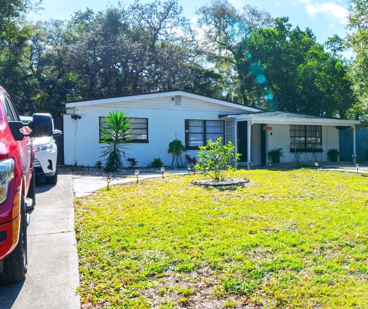 A charming white house with a spacious yard is surrounded by lush greenery. The front yard features an assortment of plants and is accentuated by a circular stone garden bed. A paved driveway is visible with parked vehicles alongside.