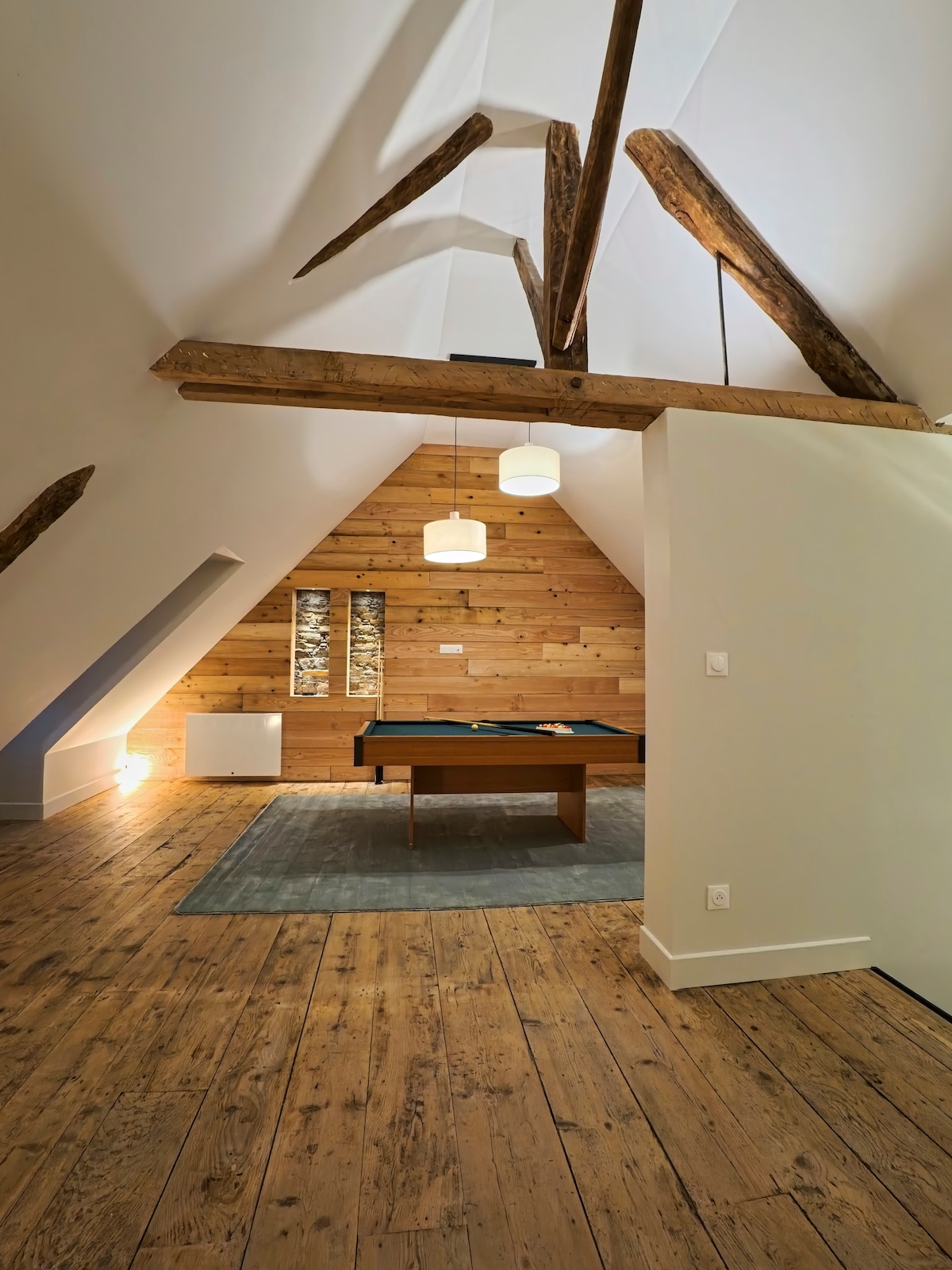 A spacious room features sloped ceilings with exposed wooden beams. A wooden table is positioned centrally on a light area rug, while warm lighting enhances the wooden walls. Large windows allow natural light to fill the space, contributing to a cozy ambiance.