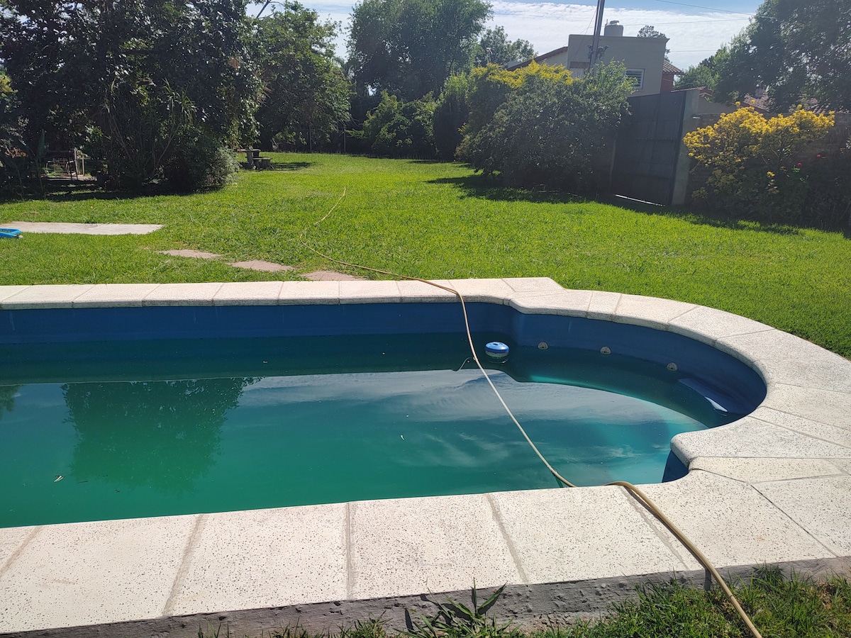 A swimming pool is situated within a spacious grassy area, surrounded by trees and vibrant greenery. The pool features a light-colored stone border, and a gentle slope leading into the water. Sunlight reflects off the water's surface, enhancing the serene outdoor setting.