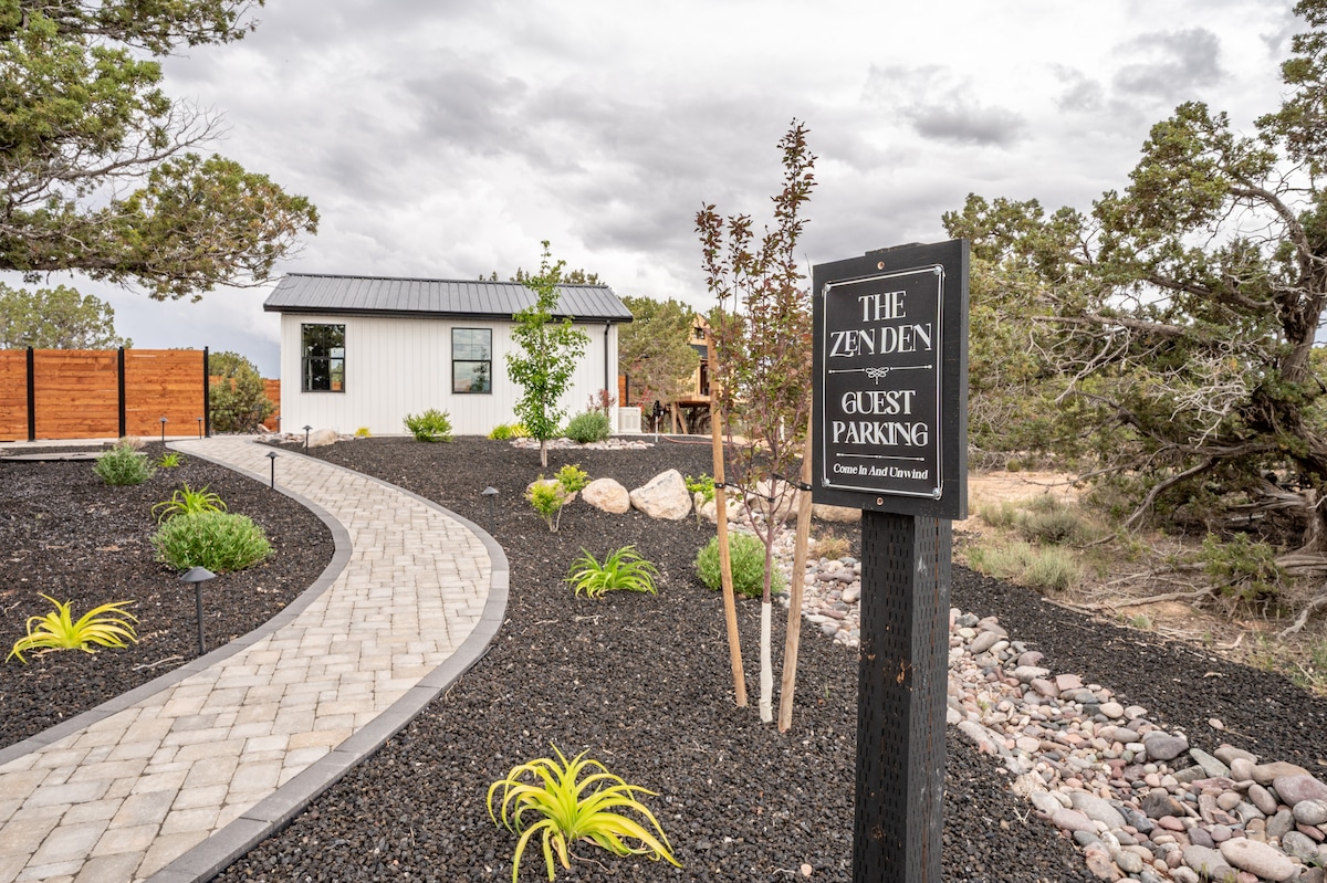 A path made of stone pavers winds through a landscaped area featuring low-maintenance plants and rocks. A sign indicating 'The Zen Den Guest Parking' stands at the forefront, guiding guests to their destination, while the serene home is visible in the background.