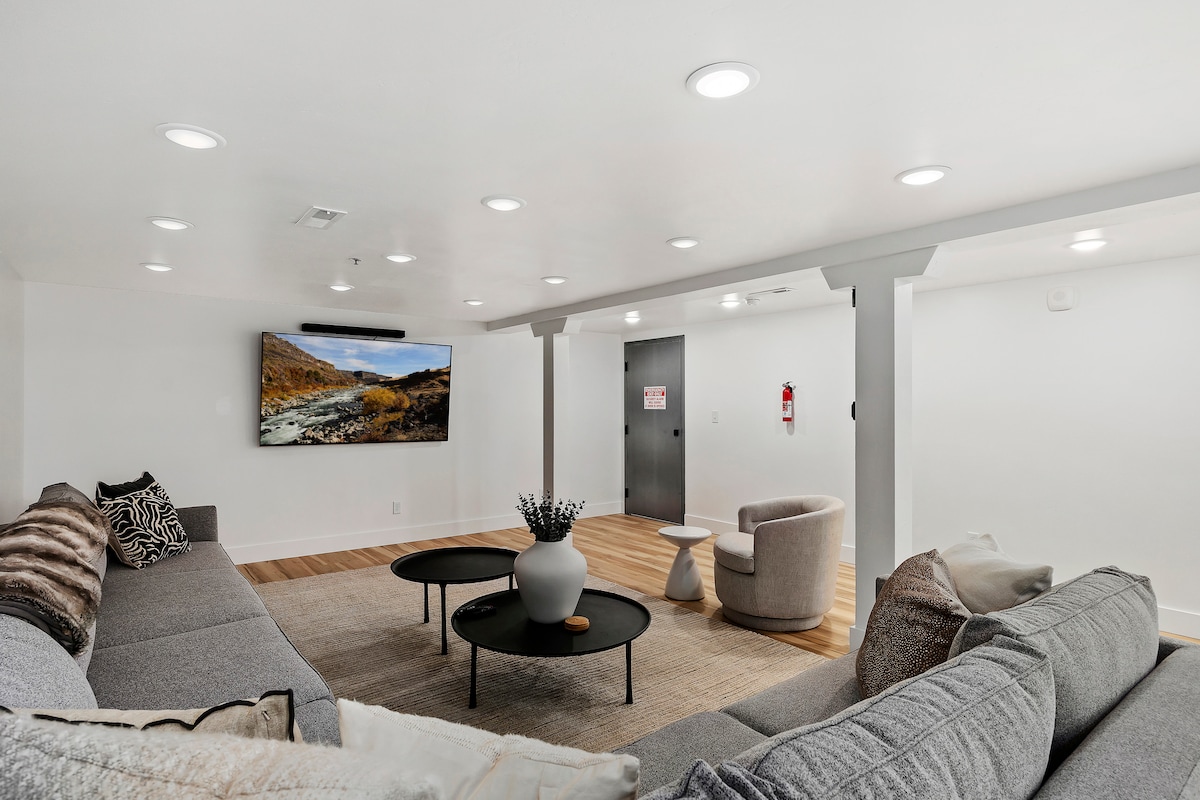 The spacious living area features a large sectional sofa and a comfortable armchair, arranged around a low coffee table. A mounted television is visible on the wall, and recessed lighting enhances the bright ambiance. Light hardwood flooring adds warmth to the modern decor.