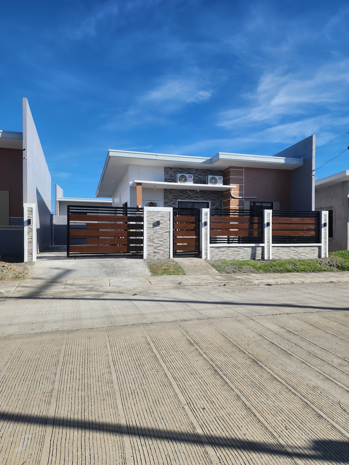 A modern, single-story home is presented with a sleek facade, featuring a combination of stone and wooden accents. A gated entrance provides security and privacy, while a clear blue sky serves as a backdrop, enhancing the inviting appearance of the property.